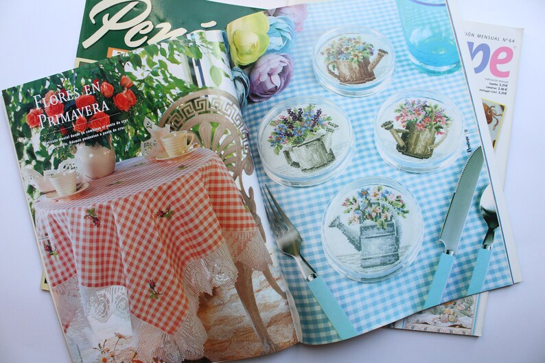 May include: A magazine spread featuring a cross-stitch pattern for a set of four coasters. Each coaster features a watering can filled with flowers. The magazine is open to a page with a blue and white checkered background and the text "ION MENSUAL N 64".