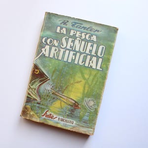 以下が含まれることがあります： R. Earlen 著、Sintés Barcelona 出版のヴィンテージブック「La Pesca Con Señuelo Artificial」。表紙には、口にルアーをくわえた魚のイラストが描かれている。