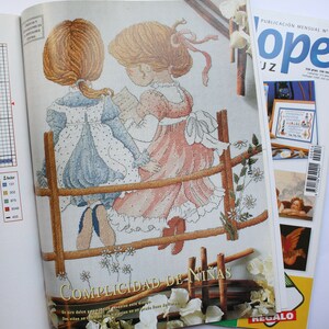 May include: A cross-stitch pattern featuring two girls in dresses standing on a wooden fence. The pattern is titled "Complicidad de Ni&ntilde;as" and includes a description in Spanish. The pattern is from a magazine called "Ope".