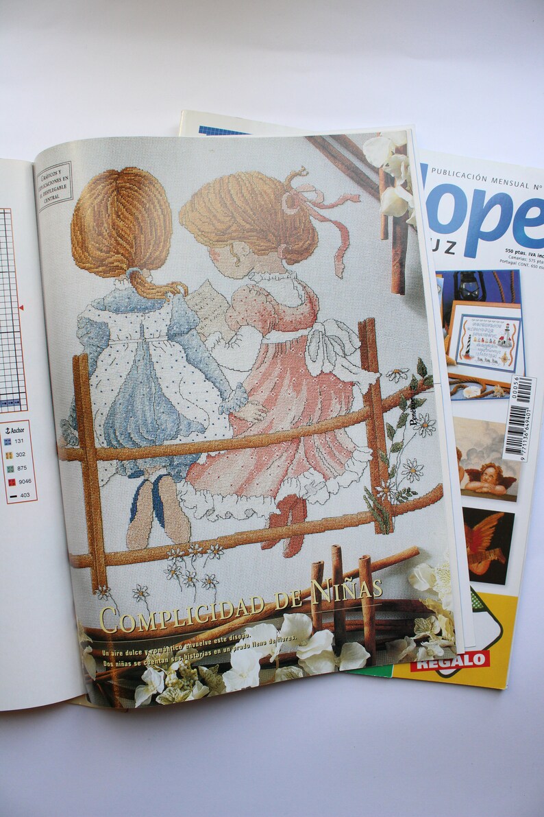 May include: A cross-stitch pattern featuring two girls in dresses standing on a wooden fence. The pattern is titled "Complicidad de Ni&ntilde;as" and includes a description in Spanish. The pattern is from a magazine called "Ope".