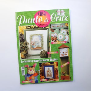 May include: A green magazine cover with the title "Casa Punto de Cruz" and images of cross-stitch designs, including a window, a clock, a bunny, and a bottle. The text "Exclusivos y espectaculares dise&ntilde;os" is at the bottom of the cover.