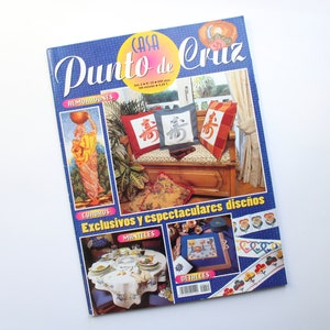 May include: A magazine cover featuring cross-stitch designs. The cover features a blue background with the title "Casa Punto de Cruz" in white lettering. The cover also features images of cross-stitch projects, including pillows, tablecloths, and borders.