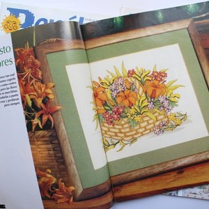 May include: A magazine page with a close-up of a framed cross-stitch design of a basket of flowers. The flowers are in shades of orange, yellow, and pink. The text on the page reads "Un cesto de flores".