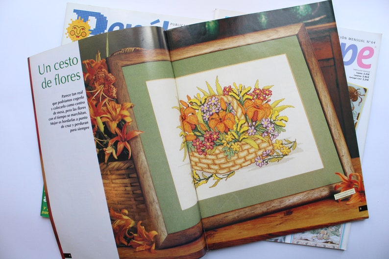 May include: A magazine page with a close-up of a framed cross-stitch design of a basket of flowers. The flowers are in shades of orange, yellow, and pink. The text on the page reads "Un cesto de flores".