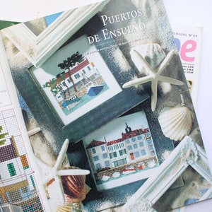 May include: A magazine cover with the title "Puertos de Ensueno" which translates to "Dream Ports" in English. The cover features two cross-stitch patterns of seaside towns framed in white wood. The background is a sandy beach with seashells and a starfish.