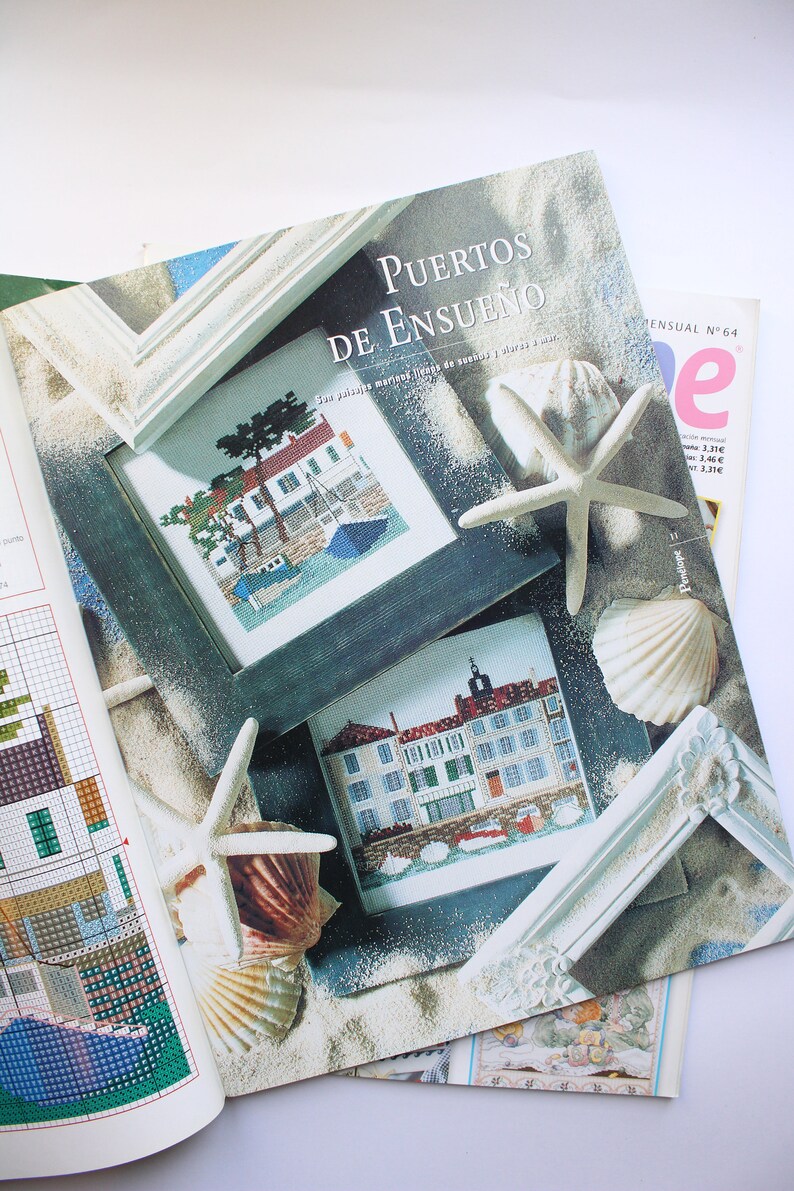 May include: A magazine cover with the title "Puertos de Ensueno" which translates to "Dream Ports" in English. The cover features two cross-stitch patterns of seaside towns framed in white wood. The background is a sandy beach with seashells and a starfish.