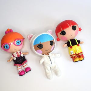 May include: Three colourful Lalaloopsy dolls with button eyes. One doll has orange hair and glasses, another has blue hair and a white cat-eared outfit, and the third has red hair and a yellow outfit.