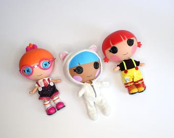 May include: Three colorful Lalaloopsy dolls with button eyes. One doll has orange hair and glasses, another has blue hair and a white cat-eared outfit, and the third has red hair and a yellow outfit.