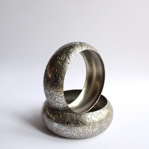 May include: Two silver-colored bangles with a floral pattern. The bangles are stacked, with one resting inside the other. The floral design is etched into the metal, creating a textured surface. The background is plain white.