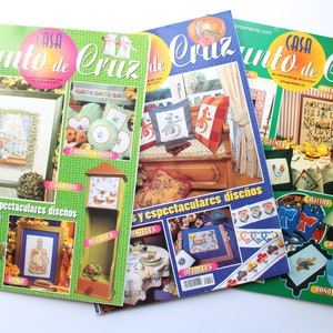 May include: Three colorful magazines with the title "Casa Punto de Cruz" on the cover. Each magazine features cross-stitch patterns and projects, including pillows, cushions, and a clock.