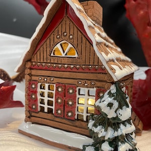 以下が含まれることがあります： 赤い屋根と白い雪が積もったセラミック製のクリスマスの家。家の前には、白い雪が積もった小さな木があります。窓は灯りがともっています。