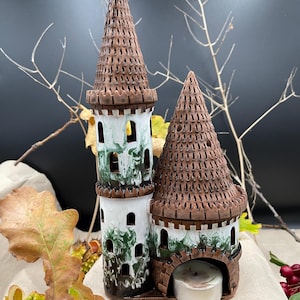 Puede incluir: Un portavelas de cerámica con forma de castillo de dos torres con techo marrón y detalles verdes. El castillo tiene una base blanca y ventanas. Una pequeña vela blanca se coloca en la abertura del castillo.