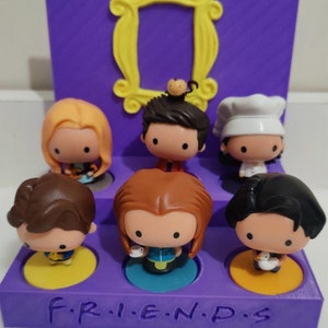 Könnte beinhalten: Ein lilafarbener Ständer mit dem Schriftzug "F.R.I.E.N.D.S." auf der Vorderseite. Der Ständer hat sechs Fächer, in denen jeweils eine andere Figur aus der Fernsehserie "Friends" im Chibi-Stil zu sehen ist. Die Figuren sind Monica, Chandler, Phoebe, Joey, Rachel und Ross.