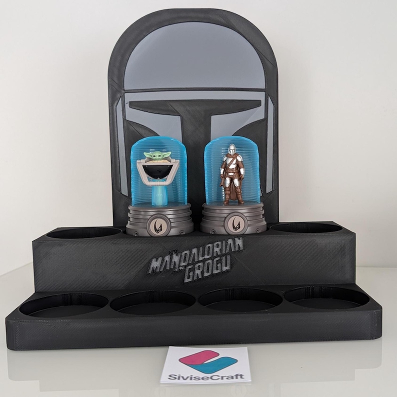 K&ouml;nnte beinhalten: Ein schwarzer Display-St&auml;nder mit einem Mandalorianer-Helm-Design. Er h&auml;lt zwei Figuren in blauen Kuppeln: Grogu und den Mandalorianer. Der St&auml;nder tr&auml;gt den Text "MANDALORIAN GROGU".