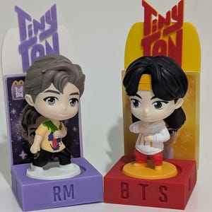 Può includere: Due statuette Tiny Tan, una su una base viola con l'etichetta "RM" e l'altra su una base rossa con l'etichetta "BTS". Le statuette sono raffigurazioni in stile cartone animato di persone, ognuna con un abbigliamento e un'acconciatura unici. La confezione presenta il logo "Tiny Tan".