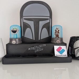 K&ouml;nnte beinhalten: Eine Pr&auml;sentation von Star Wars-Artikeln. Enth&auml;lt zwei Tassen, eine wei&szlig;e mit Grogu, die andere schwarze mit einem Mandalorianer-Helm-Design. Au&szlig;erdem ein schwarzer Pr&auml;sentationsst&auml;nder mit Mandalorianer-Helm-Kunst und zwei Figuren.