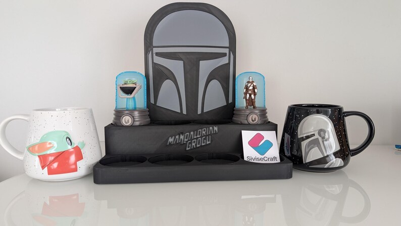 K&ouml;nnte beinhalten: Eine Pr&auml;sentation von Star Wars-Artikeln. Enth&auml;lt zwei Tassen, eine wei&szlig;e mit Grogu, die andere schwarze mit einem Mandalorianer-Helm-Design. Au&szlig;erdem ein schwarzer Pr&auml;sentationsst&auml;nder mit Mandalorianer-Helm-Kunst und zwei Figuren.