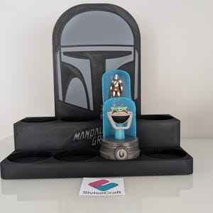 K&ouml;nnte beinhalten: Ein schwarzer Mandalorian-Themen-Display-St&auml;nder mit Helm-Design und dem Text "Mandalorian Grogu". Er enth&auml;lt eine Vitrine mit Grogu- und Mandalorian-Figuren. Der St&auml;nder hat mehrere kreisf&ouml;rmige Aussparungen.