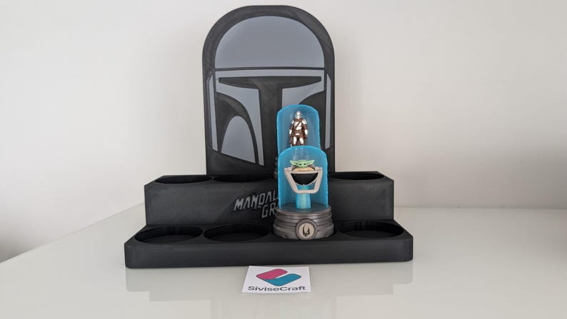 K&ouml;nnte beinhalten: Ein schwarzer Mandalorian-Themen-Display-St&auml;nder mit Helm-Design und dem Text "Mandalorian Grogu". Er enth&auml;lt eine Vitrine mit Grogu- und Mandalorian-Figuren. Der St&auml;nder hat mehrere kreisf&ouml;rmige Aussparungen.