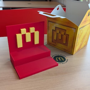 Puede incluir: Una caja de cartón roja y amarilla con un logotipo M estilizado. La caja está sentada en una mesa de madera. El logotipo está hecho de bloques pixelados.