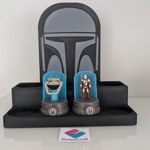 K&ouml;nnte beinhalten: Ein schwarzer Mandalorian-Themen-Display-St&auml;nder mit zwei Figurenhaltern, einer mit einer Grogu-Figur und der andere mit einer Mandalorian-Figur. Der St&auml;nder hat ein Helmdesign auf der R&uuml;ckseite und ein Logo unten.