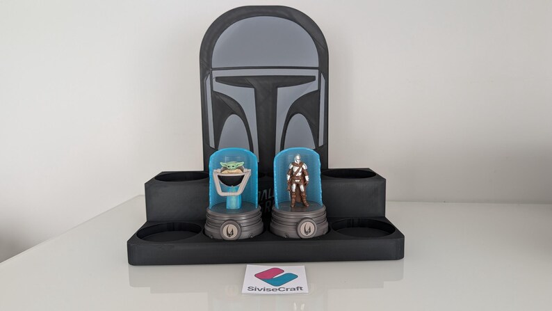 K&ouml;nnte beinhalten: Ein schwarzer Mandalorian-Themen-Display-St&auml;nder mit zwei Figurenhaltern, einer mit einer Grogu-Figur und der andere mit einer Mandalorian-Figur. Der St&auml;nder hat ein Helmdesign auf der R&uuml;ckseite und ein Logo unten.