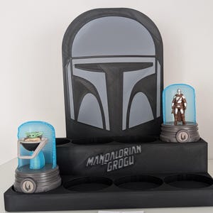 K&ouml;nnte beinhalten: Ein schwarzer Display-St&auml;nder mit einem Mandalorianer-Helm-Design. Er verf&uuml;gt &uuml;ber zwei beleuchtete Vitrinen mit Figuren von Grogu und dem Mandalorianer. Auf dem St&auml;nder steht "Mandalorian Grogu".