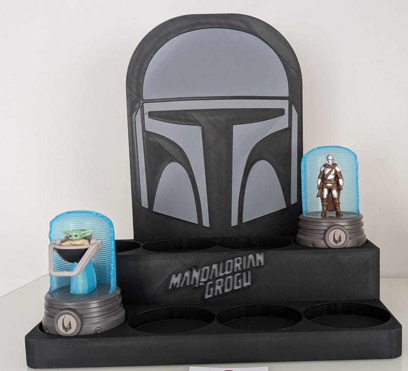 K&ouml;nnte beinhalten: Ein schwarzer Display-St&auml;nder mit einem Mandalorianer-Helm-Design. Er verf&uuml;gt &uuml;ber zwei beleuchtete Vitrinen mit Figuren von Grogu und dem Mandalorianer. Auf dem St&auml;nder steht "Mandalorian Grogu".