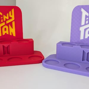 Può includere: Espositori Tiny Tan rossi e viola. Ogni espositore ha una base rettangolare con ritagli circolari, una sezione centrale con tre scomparti e un pannello posteriore con il logo Tiny Tan. L'espositore rosso ha testo giallo, quello viola ha testo bianco.