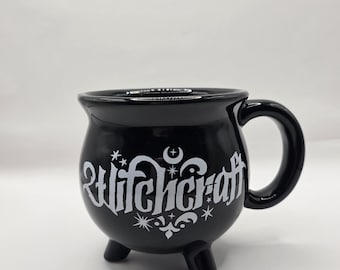 May include: Black ceramic cauldron-shaped mug with a handle and three small feet. The mug features the word "Witchcraft" in white stylized lettering, along with star and crescent moon designs. The mug is designed for beverages.