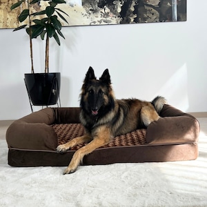 Op de afbeelding: Een bruin hondenbed met een pluche, bruine buitenkant van stof en een geruite bruine en beige binnenvoering. Een bruine en zwarte hond ligt op het bed.