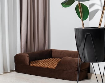 Può includere: Un letto per cani marrone con un bordo rialzato e un cuscino con motivo marrone e beige. Il letto si trova in una stanza con tende bianche e marroni e una pianta in vaso.
