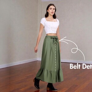 Op de afbeelding: Een vrouw draagt een witte crop top met vierkante hals en een olijfgroene maxi rok. De rok heeft een knoopsluiting aan de voorkant, een strikceintuur en een gefranjerde zoom. Zwarte enkellaarsjes maken de outfit compleet.