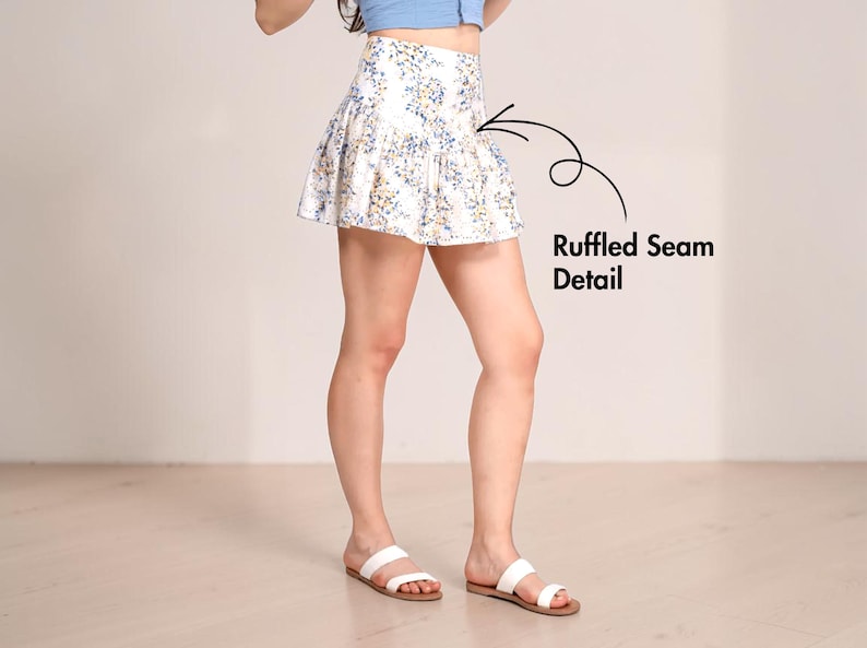 May include: A white ruffled mini skirt with a floral pattern in blue and yellow. The skirt has a high waist and a flared silhouette. The image also shows white sandals.