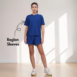Puede incluir: Una mujer con un conjunto de camiseta y pantal&oacute;n corto azul a juego, con mangas ragl&aacute;n. La camiseta tiene cuello redondo y mangas cortas. El pantal&oacute;n corto es de un tono azul similar. Lleva zapatillas blancas. El texto "Raglan Sleeves" est&aacute; en la imagen.