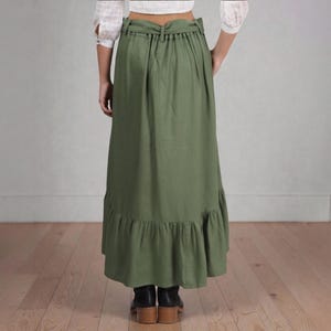 Op de afbeelding: Olijfgroene maxi rok met een geplooid zoom. De rok heeft een hoge taille en een trekkoord detail. Het model draagt zwarte laarzen. De rok is gemaakt van een lichte stof.