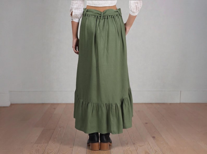 Op de afbeelding: Olijfgroene maxi rok met een geplooid zoom. De rok heeft een hoge taille en een trekkoord detail. Het model draagt zwarte laarzen. De rok is gemaakt van een lichte stof.