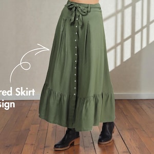 Op de afbeelding: Olijfgroene rok met lagen, een knoopsluiting en een strik in de taille. De rok heeft een geplooid zoom en wordt gecombineerd met zwarte enkellaarsjes. De tekst "Tiered Skirt Design" is zichtbaar.