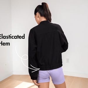 Pode incluir: &Eacute; apresentada uma jaqueta preta com bainha el&aacute;stica. A modelo est&aacute; usando shorts lil&aacute;s. O texto "Bainha el&aacute;stica" &eacute; exibido no lado esquerdo da imagem.