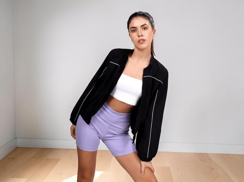 Pode incluir: Uma jaqueta bomber preta com acabamento branco &eacute; usada sobre um top de tubo branco e shorts de ciclista lavanda. A jaqueta tem gola redonda e mangas compridas. A modelo est&aacute; em p&eacute; em um piso de madeira.