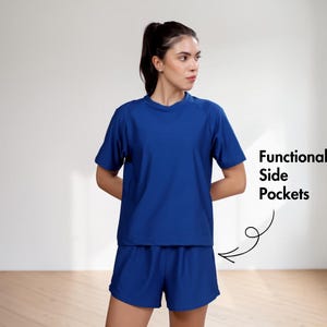 Puede incluir: Una mujer con un conjunto deportivo azul a juego de camiseta y pantal&oacute;n corto. La camiseta tiene mangas cortas y cuello redondo. El pantal&oacute;n corto tiene cintura el&aacute;stica. El texto en la imagen dice "Functional Side Pockets".