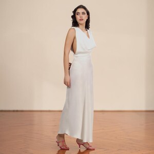 May include: A sleeveless, floor-length white dress with a cowl neckline. The dress has a flowing silhouette and is paired with pink heeled sandals. The background is a neutral beige, and the floor is a light brown wood.
