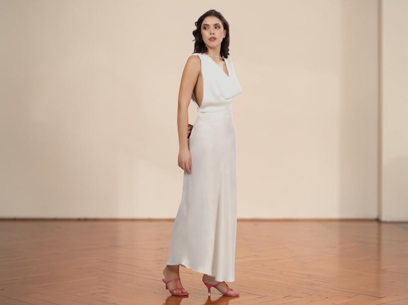 May include: A sleeveless, floor-length white dress with a cowl neckline. The dress has a flowing silhouette and is paired with pink heeled sandals. The background is a neutral beige, and the floor is a light brown wood.