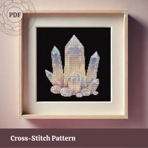 May include: A framed cross-stitch pattern featuring a cluster of crystals in shades of cream, lavender, and blue against a black background. The artwork is displayed in a light wood frame with a white mat. The text "Cross-Stitch Pattern" is at the bottom.