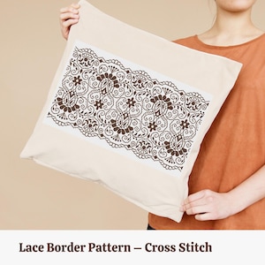 May include: A cream-coloured cushion featuring a cross-stitch lace border design in brown. The cushion is held up by a person. The text at the bottom of the image reads "Lace Border Pattern - Cross Stitch".