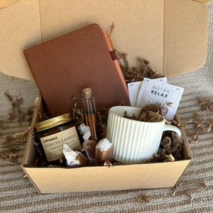 May include: A brown cardboard box filled with a self-care gift set. The box contains a brown leather journal, a white mug, a small glass jar of scented candles, a small glass bottle with matches, and a few caramel candies. The box is filled with brown packing paper.
