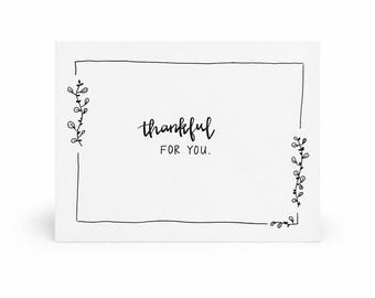 May include: A white greeting card with the words "thankful FOR YOU" in black script. The card has a black border with small leaf designs in each corner and along the sides.