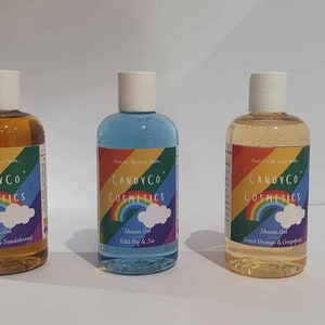 Puede incluir: Tres botellas de gel de ducha transparentes con tapas blancas. Cada botella tiene una etiqueta con el texto "CANDYCO COSMETICS", un arcoíris y una nube. Las botellas contienen gel de ducha en colores ámbar, azul y amarillo. Las etiquetas dicen "Black Pepper & Sandalwood", "Wild Fig & Iris" y "Sweet Orange & Grapefruit".