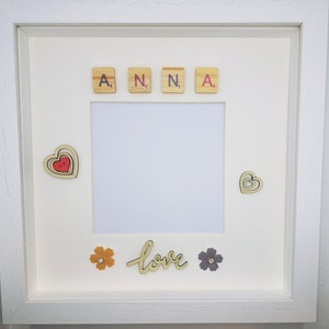 May include: A white picture frame with a white mount and a white background. The frame has a cutout in the centre for a photo. The cutout is decorated with wooden letter tiles spelling "ANNA" and a wooden cutout of the word "love". There are also wooden cutouts of hearts and flowers.