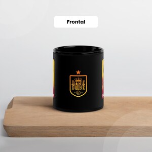 May include: Black ceramic mug with the Spanish coat of arms in gold and red. The mug has a star above the emblem and the word "Frontal" in white. The mug is on a wooden surface.